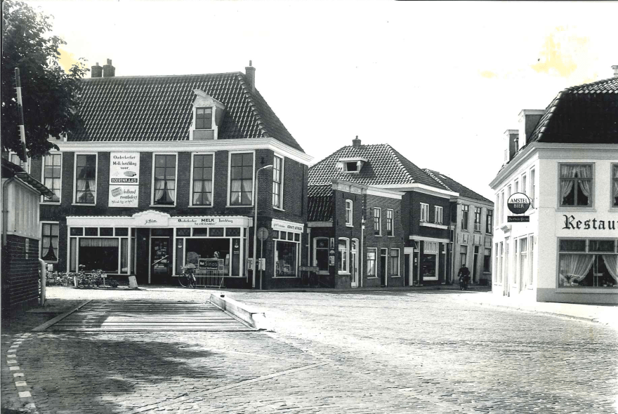 Deze foto is genomen aan de Amstelzijde in augustus 1964. Links de voertuigenweegbrug waarop landbouwwerktuigen met lading werden gewogen en bijbehorend dienstgebouwtje annex urinoir. Daarachter het winkelwoonhuis uit de 19e eeuw, waar in 1964 de Ouderkerker Melkinrichting zat van J. Blom. Rechts restaurant de Oude Prins, waar momenteel restaurant Loetje aan de Amstel in zit. De Amstelzijde met aangrenzende bebouwing is door het Rijk aangewezen als beschermd dorpsgezicht. 